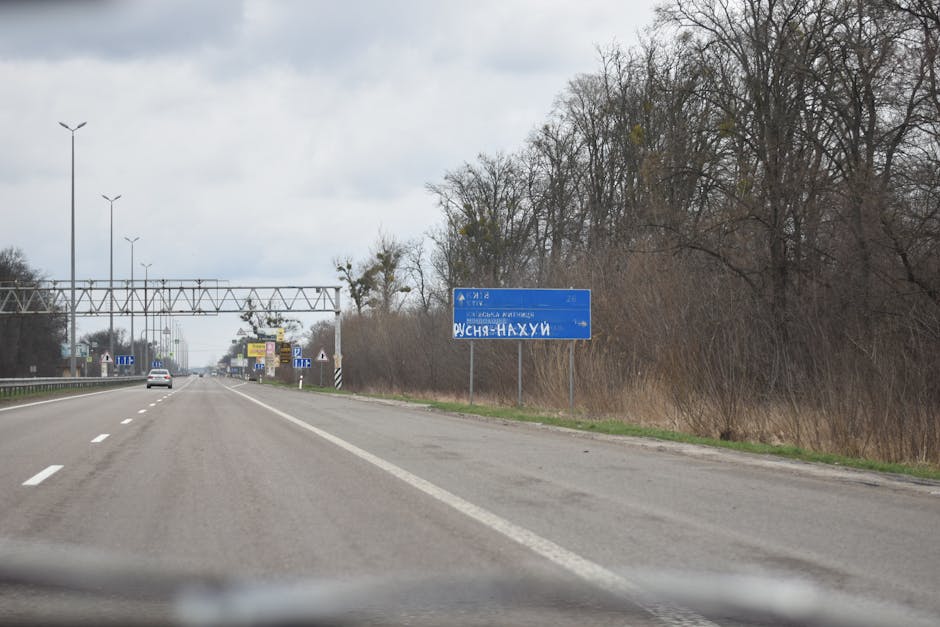 Vandalized road sign on a scenic highway surrounded by bare trees and cloudy sky.