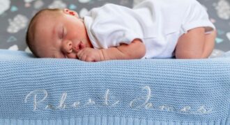 A baby sleeps peacefully on a blue knit blanket with name embroidery.