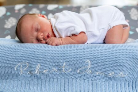 A baby sleeps peacefully on a blue knit blanket with name embroidery.