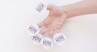 A hand displays several 'Vote' stickers on a white background, symbolizing political participation.