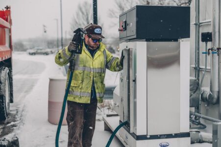 A man wearing a jacket refuels a truck at a gas station during winter with snow falling.