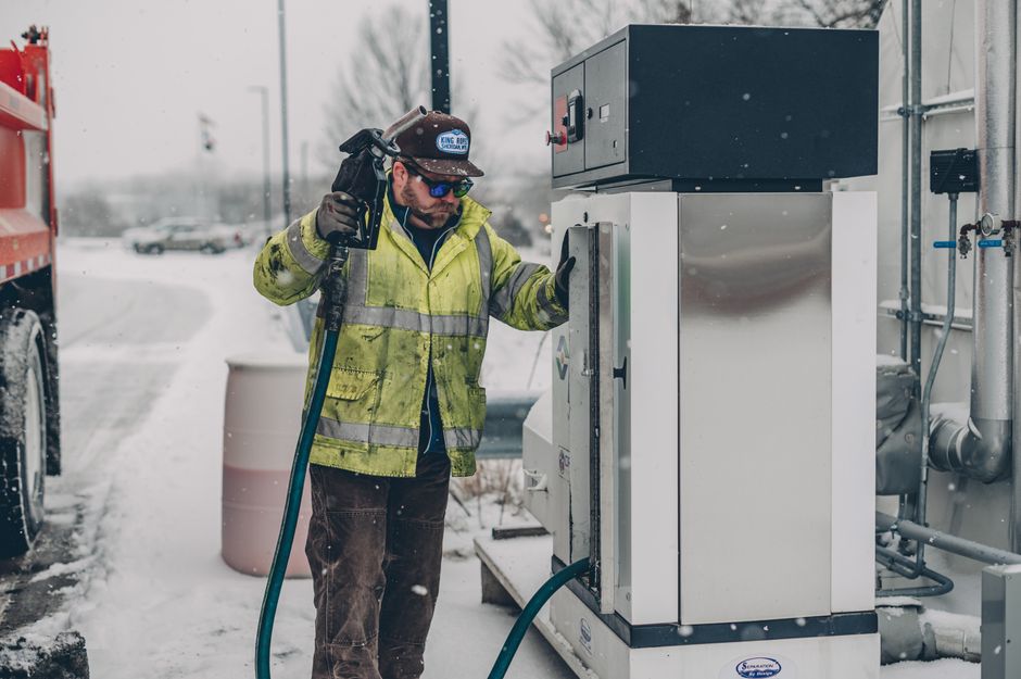 A man wearing a jacket refuels a truck at a gas station during winter with snow falling.