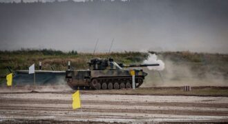 A military tank firing a shell during training in an open field.