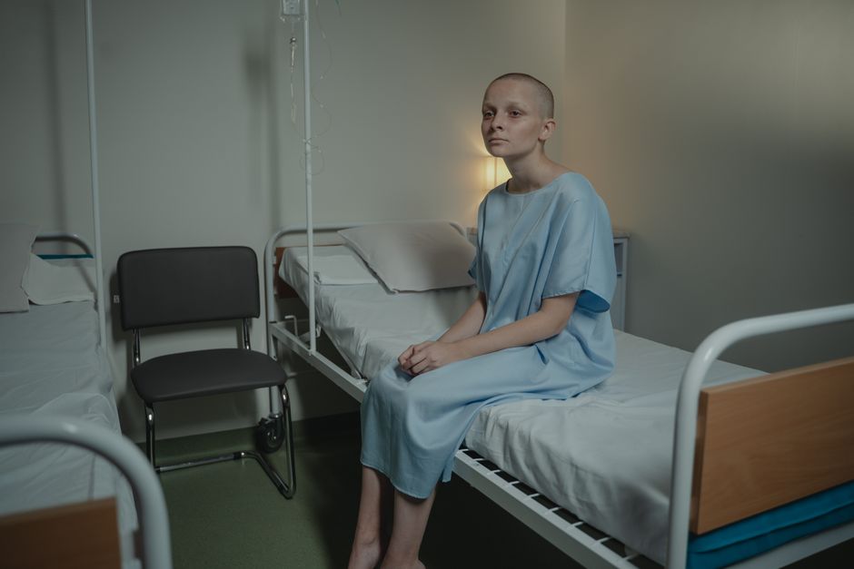 A young woman in a hospital gown sits on a bed in a medical setting, conveying a somber mood.