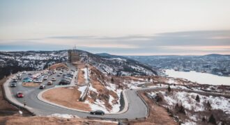 Automobiles riding on curvy asphalt roadway near grassy shore covered with snow against river and cloudy sky on winter day