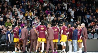 Basketball team huddles on court during a game timeout with audience in background.