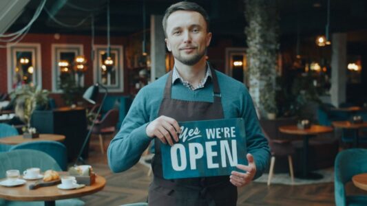 Cafe owner standing inside, holding a 'We're Open' sign, welcoming customers.