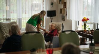 Caregiver assisting elderly women with tea in a cozy nursing home setting.