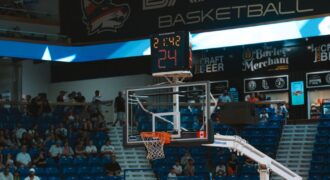 Close-up of a basketball hoop inside a stadium with a scoreboard and audience in the background.