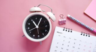 Flat lay of a pink-themed workspace featuring an alarm clock, calendar, pen, and paper clip on a pastel surface.