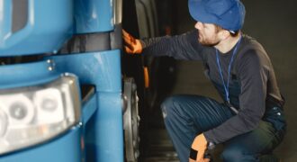 Mechanic in uniform checking a blue truck's wheel inside a garage workshop.