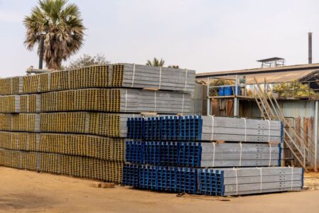 Piles of stacked metal beams in an industrial outdoor yard under a clear sky.