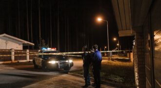 Police officers at a nighttime crime scene with a police car and caution tape under streetlights.