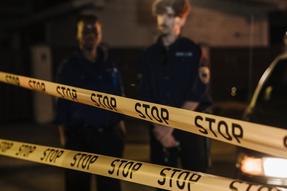 Two police officers standing behind a stop tape at night, scene partially illuminated by car headlights.