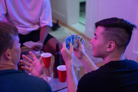 Young adults playing a card game with drinks and laughter in a cozy indoor setting.
