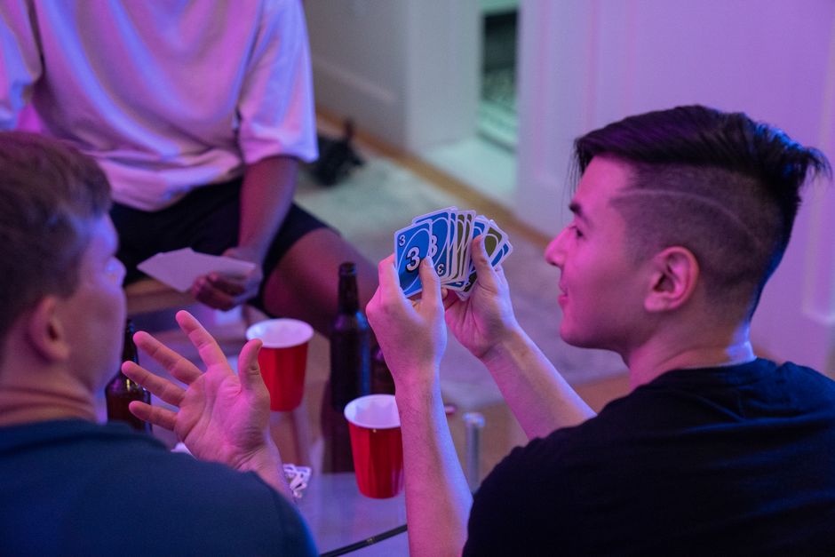 Young adults playing a card game with drinks and laughter in a cozy indoor setting.