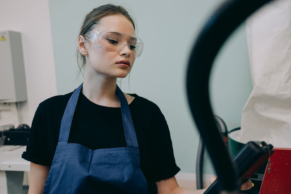 Young woman in a workshop setting, wearing protective goggles and a blue apron, standing indoors.
