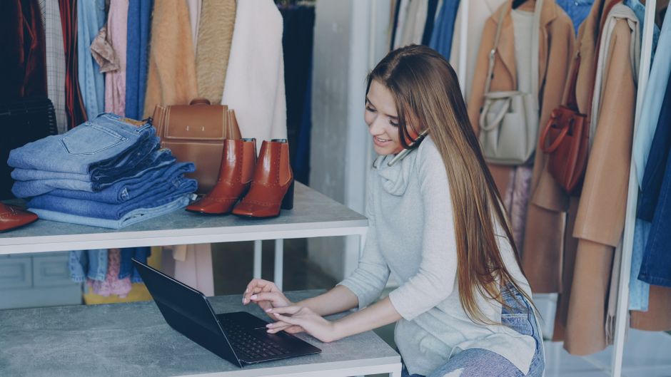 Young woman managing a fashion boutique, multitasking with phone and laptop, surrounded by clothing.